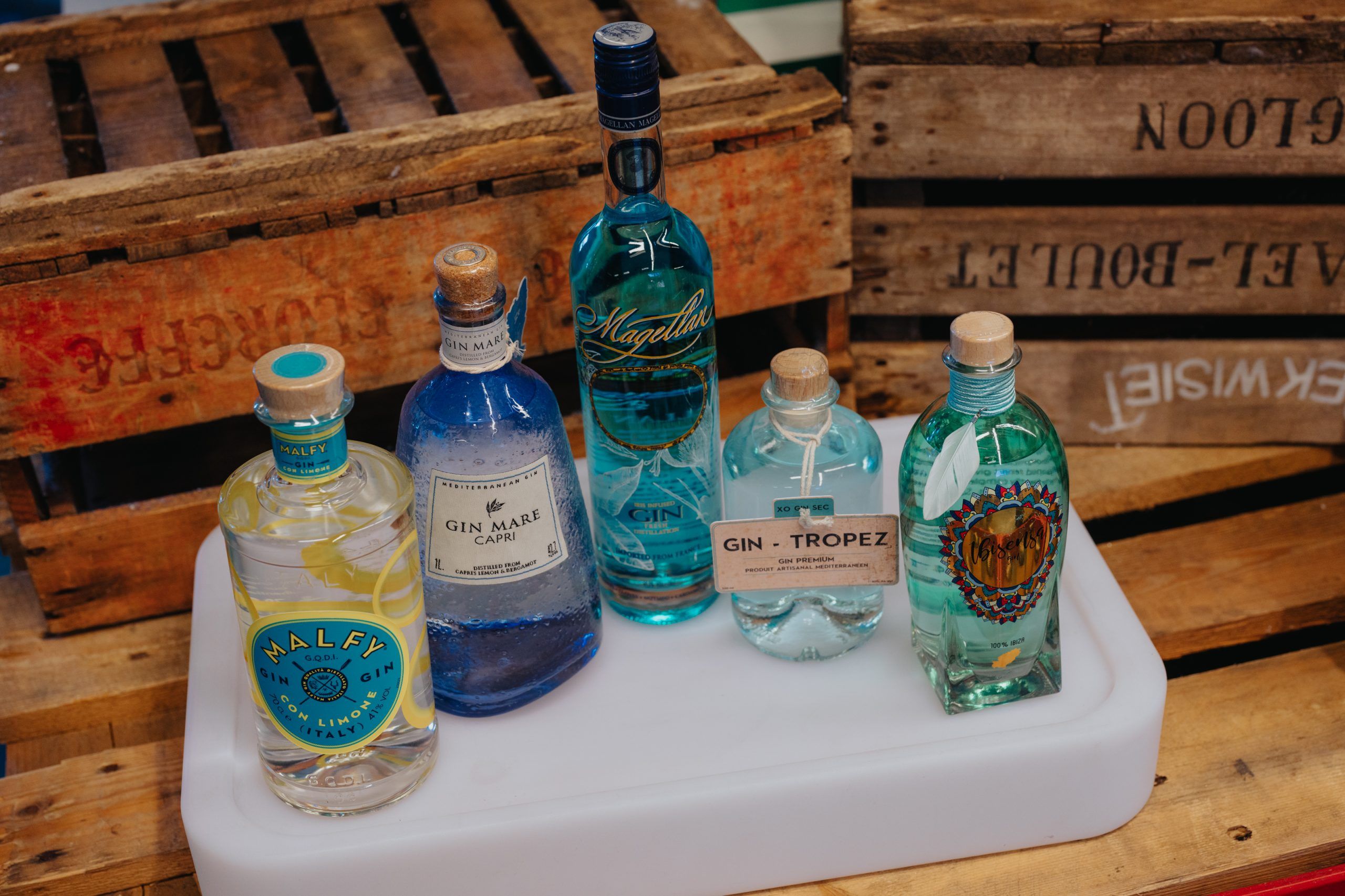 Vijf flessen gin, waaronder Malfy, Gin Mare Capri en Magellan, uitgestald op houten kisten.